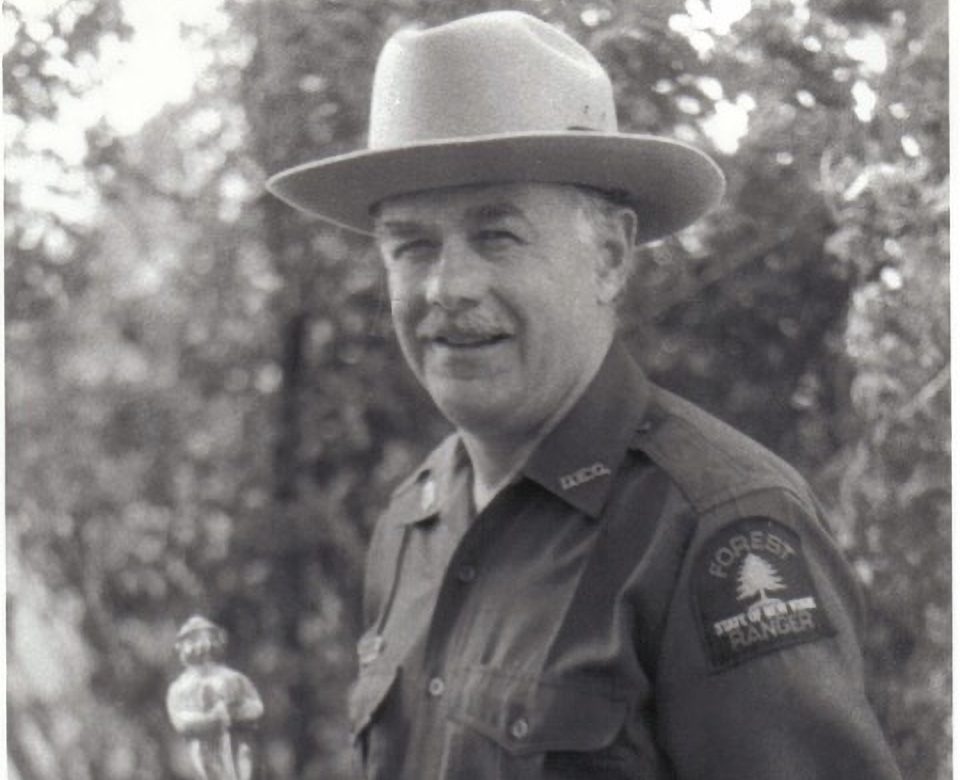 black and white photo of man in forest ranger uniform holding Smokey Bear trophy